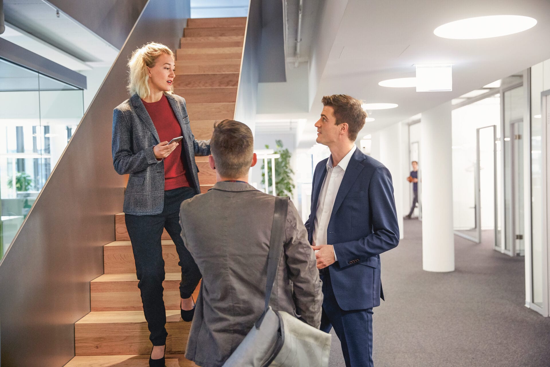 KMU-Versicherung: Frau auf einer Treppe im modernen Bürogebäude im Gespräch mit zwei Arbeitskollegen im Business-Outfit. 