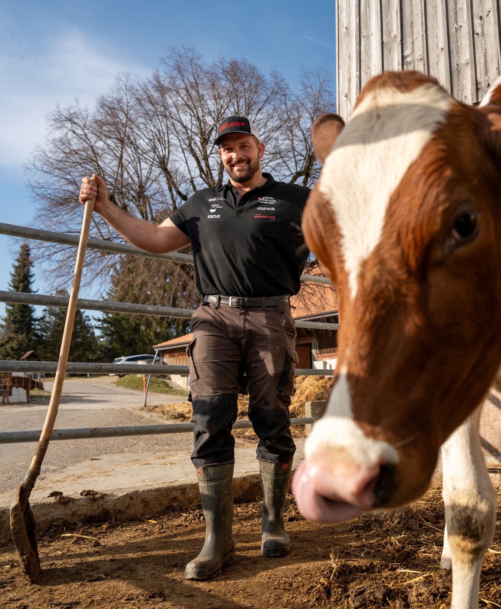Benjamin Gapany dans une étable, souriant avec une fourche à la main et une vache au premier plan – entre force et tradition.