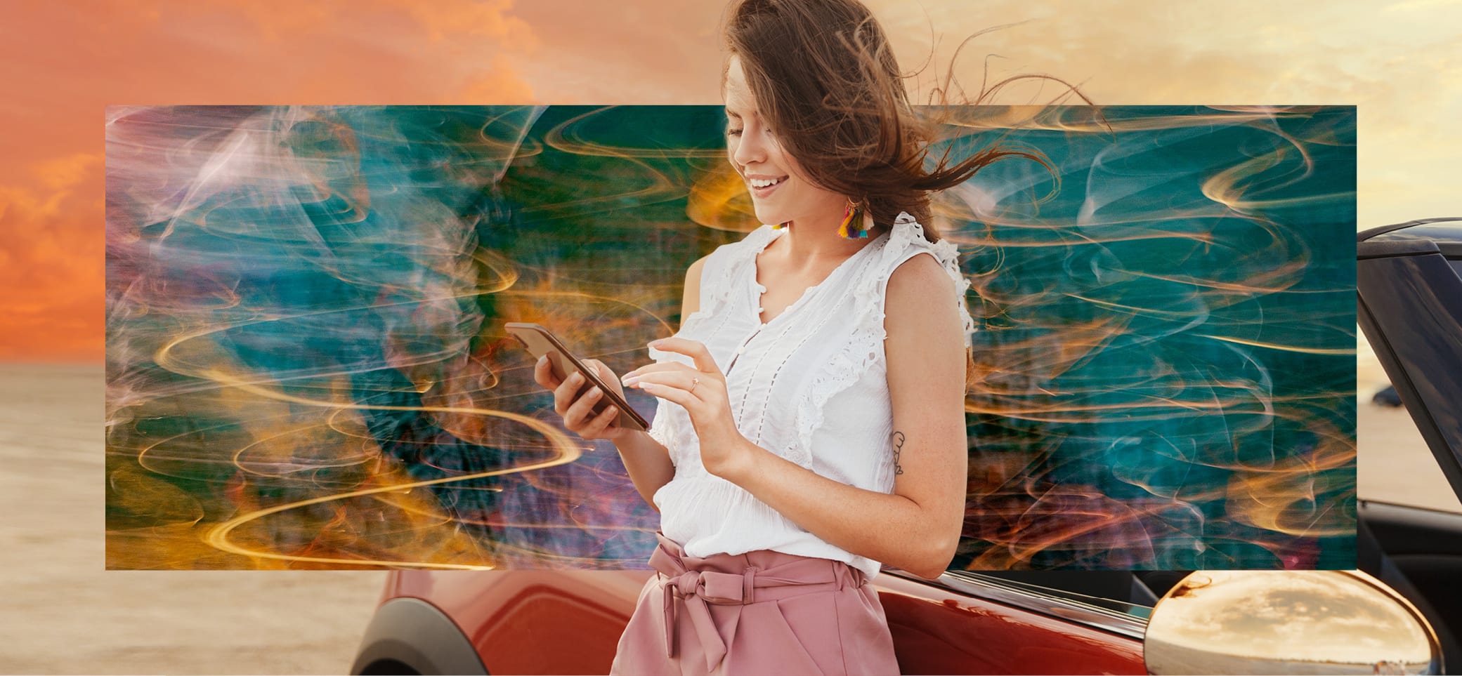 Junge Frau steht lehnt sich an ein Auto. Sie hat Wind in den Haaren und drückt lachend mit beiden Händen auf ihrem Smartphone.