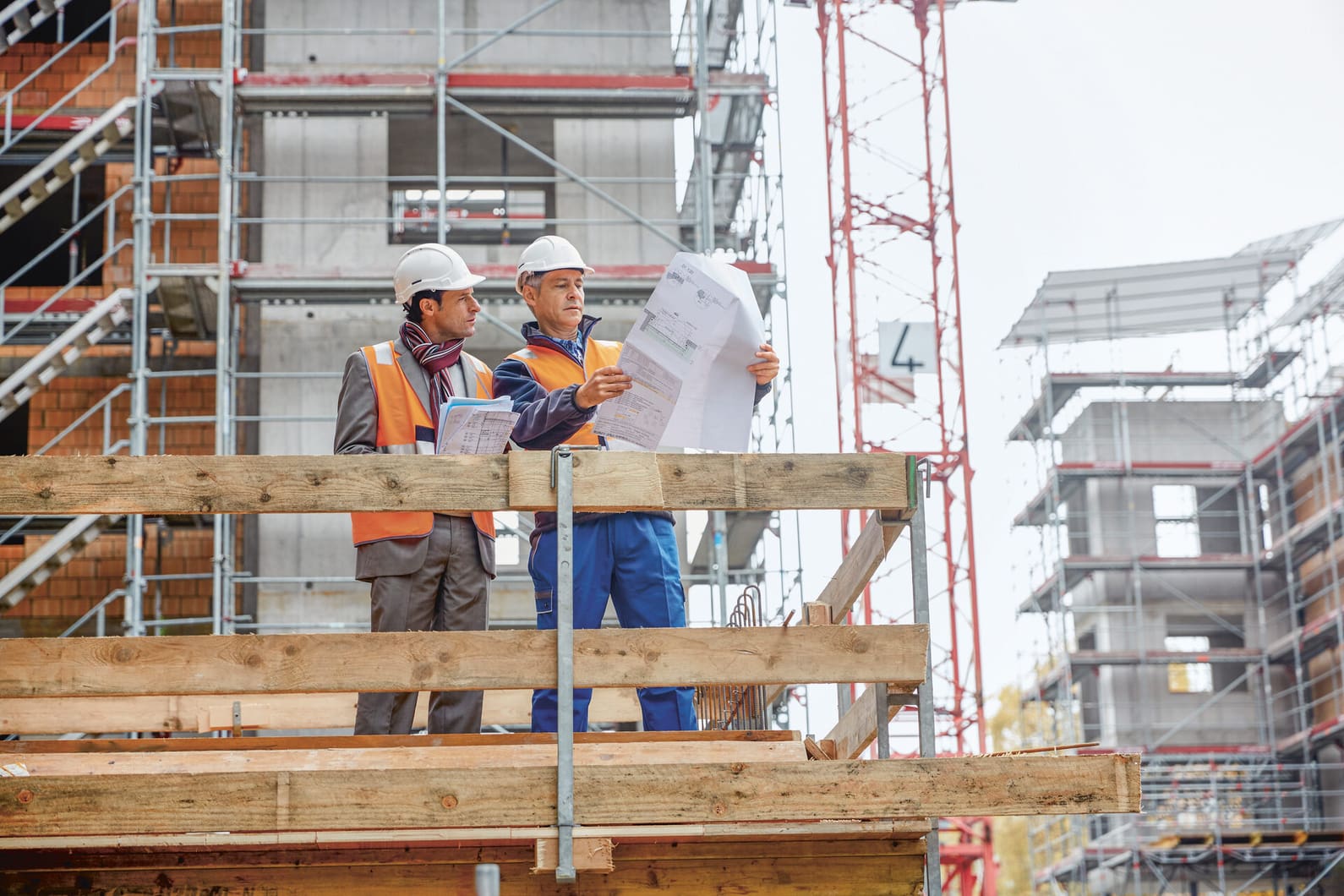 Zwei Männer mit Bauhelmen stehen auf einer Baustelle und sehen sich gemeinsam die Baupläne an. 