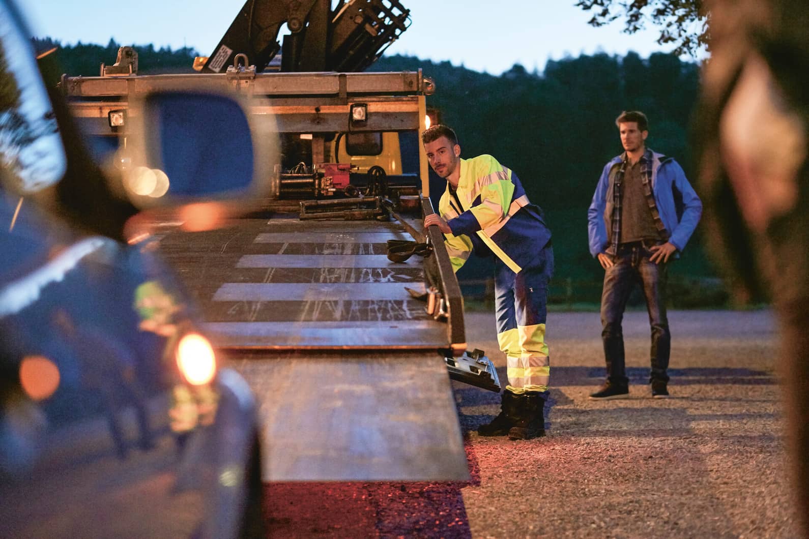 L’employé d’une entreprise de dépannage charge un véhicule visiblement en panne. Debout à côté de lui, le détenteur du véhicule assiste à la scène.