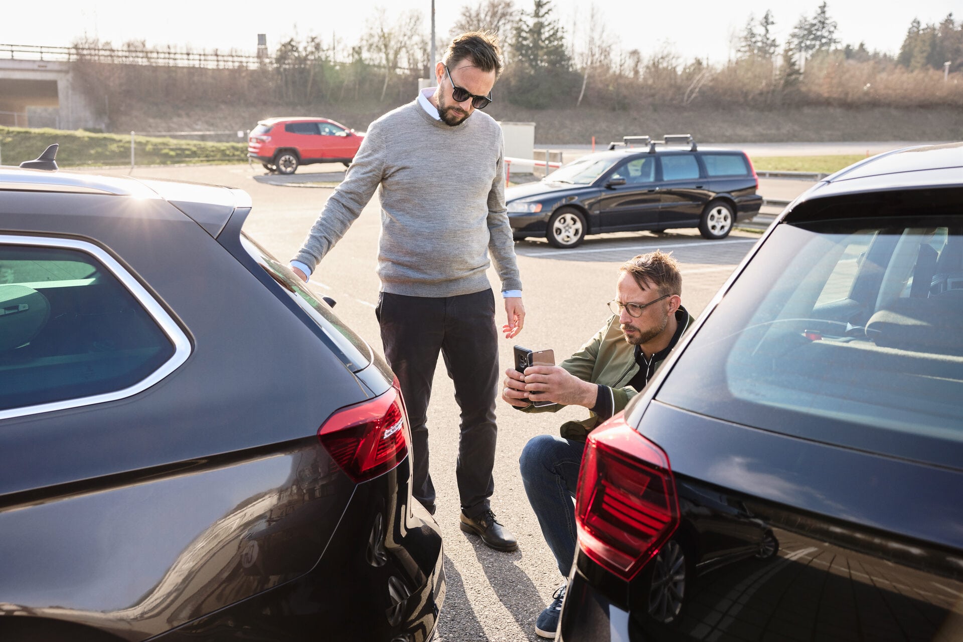 Un uomo si inginocchia accanto a un’auto e fotografa il danno dopo un incidente. Vicino c’è un’altra persona in piedi.