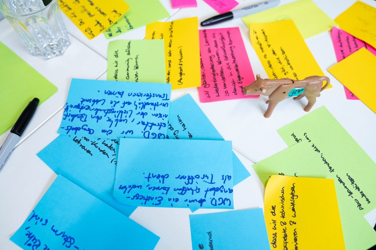 Bunte Post-its mit handschriftlichen Notizen liegen auf einem weißen Tisch, daneben ein Glas Wasser, ein Filzstift und eine kleine Plastik-Kuh mit der Aufschrift „CHF“ – Szene eines kreativen Brainstormings oder Workshops.